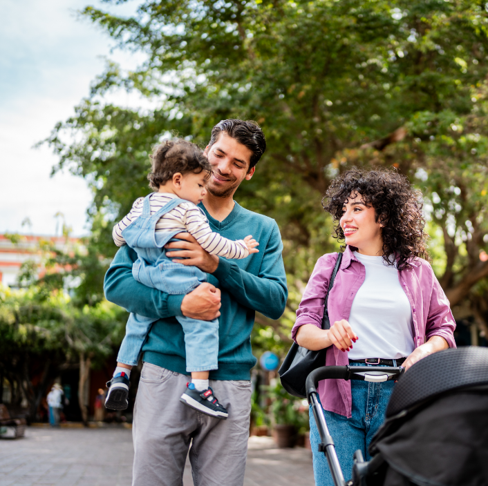 Eine Familie mit einem kleinen Kind in einem Park, der Vater hält das Kind, während die Mutter mit einem Kinderwagen steht.