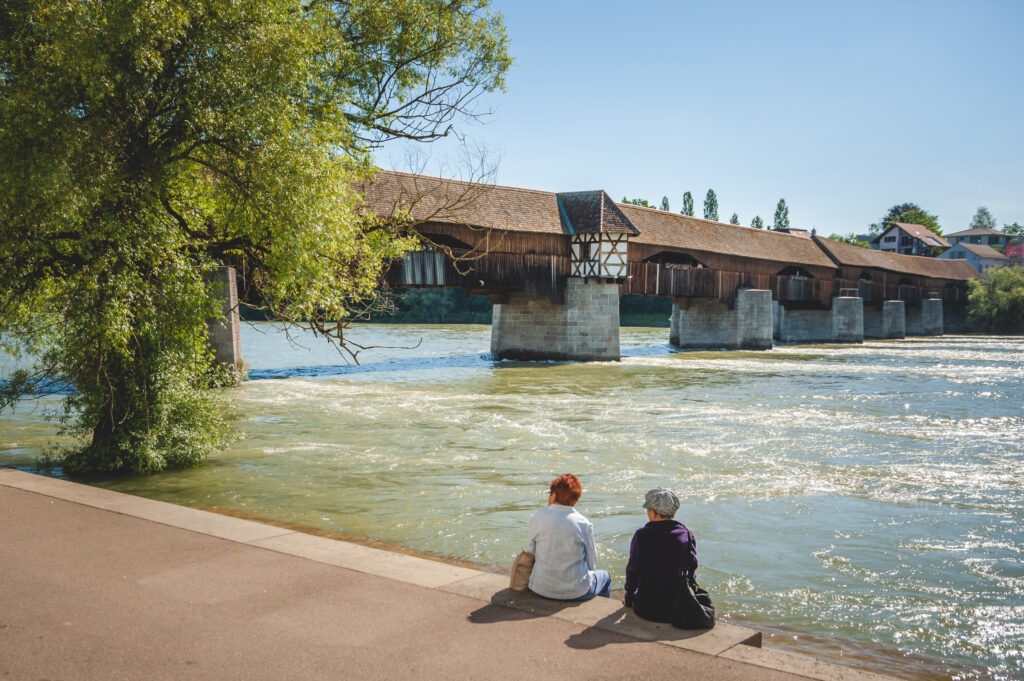 Zwei Personen sitzen am Flussufer und betrachten eine Holzbrücke, umgeben von grünen Bäumen und klarem Himmel.