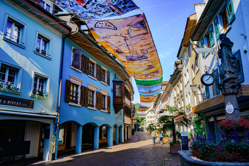 Bunte Stoffbanner hängen über einer malerischen Straße mit historischen Gebäuden und Blumenarrangements.