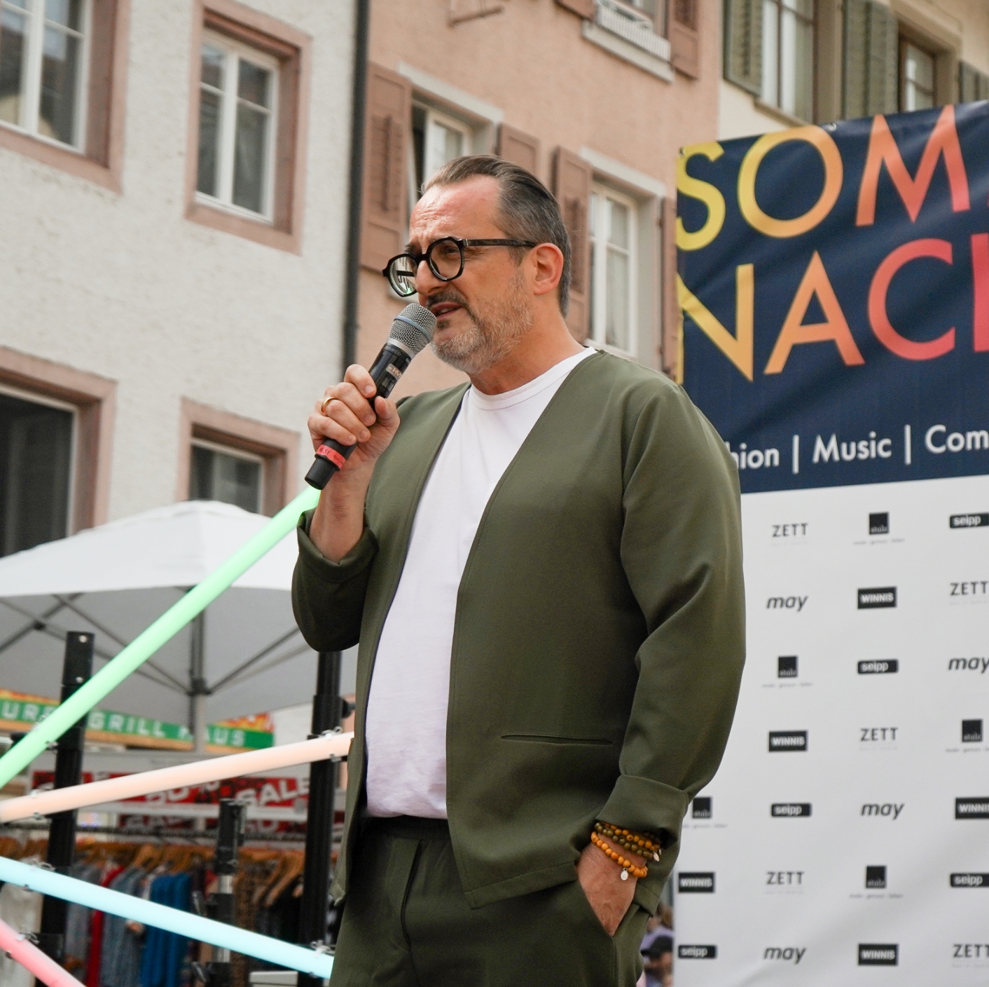 Mann mit Brille spricht ins Mikrofon auf einer Bühne, umgeben von Werbebannern und farbigen Lichtern.