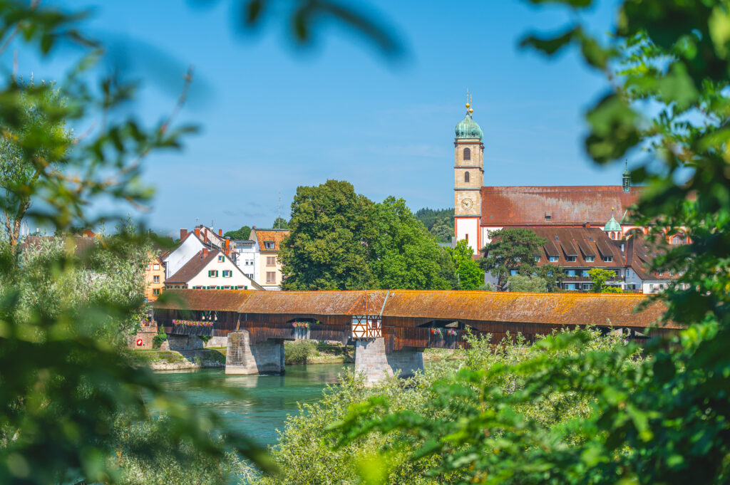 Blick auf eine Holzbrücke über einen Fluss, umgeben von Bäumen und historischen Gebäuden unter blauem Himmel.