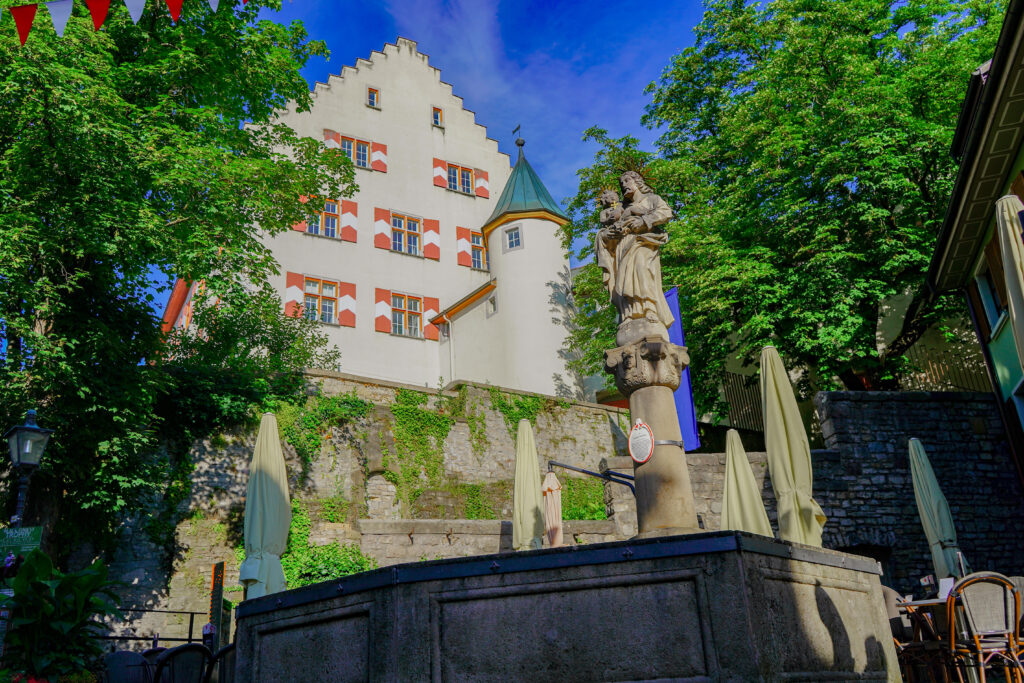 Historisches Gebäude im Hintergrund, Statue in der Mitte, umgeben von Bäumen und schattigen Gastronomietischen.