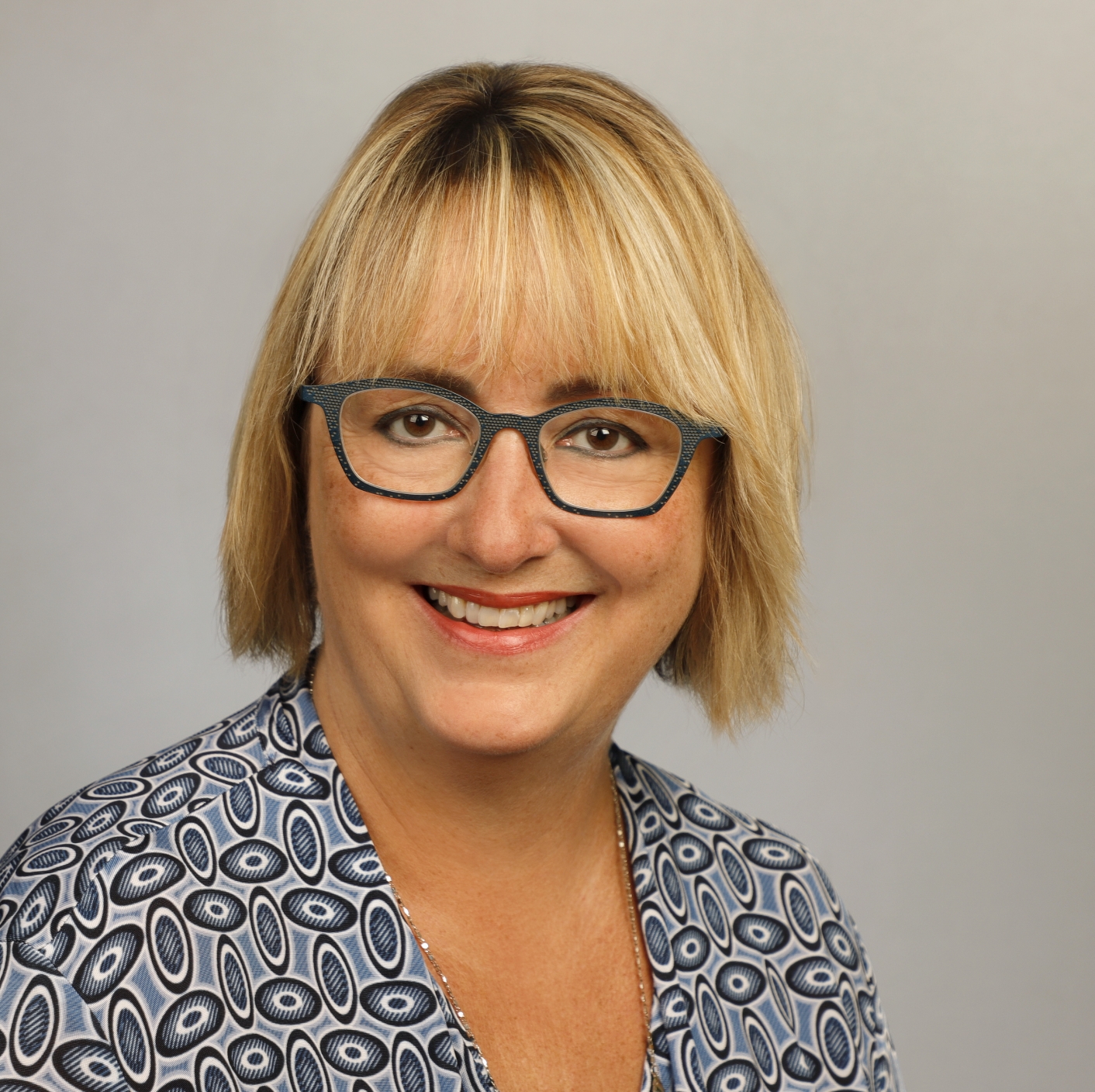 Porträt einer lächelnden Frau mit blonden, schulterlangen Haaren und Brille, die ein gemustertes Oberteil trägt.