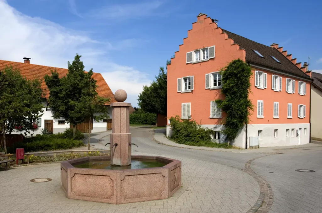 Fountain in a village square, surrounded by green trees and characterized by traditional houses and a blue sky.