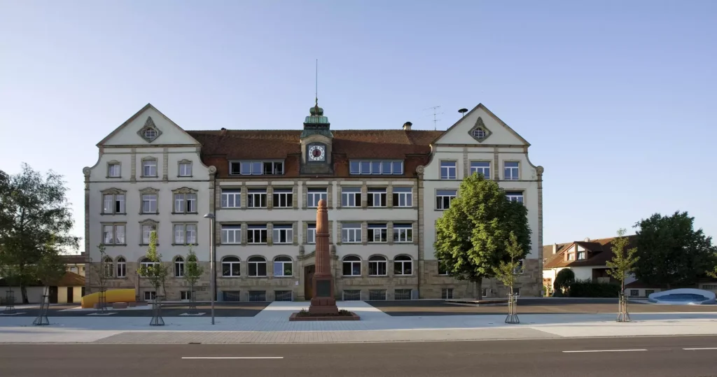 Historisches Gebäude mit Uhr, umgeben von Bäumen und einer Platzgestaltung, im Vordergrund ein Denkmal.