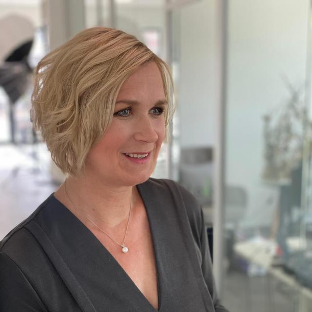 Frau mit blonden, kurz gestuften Haaren, lächelt in einem modern eingerichteten Raum.